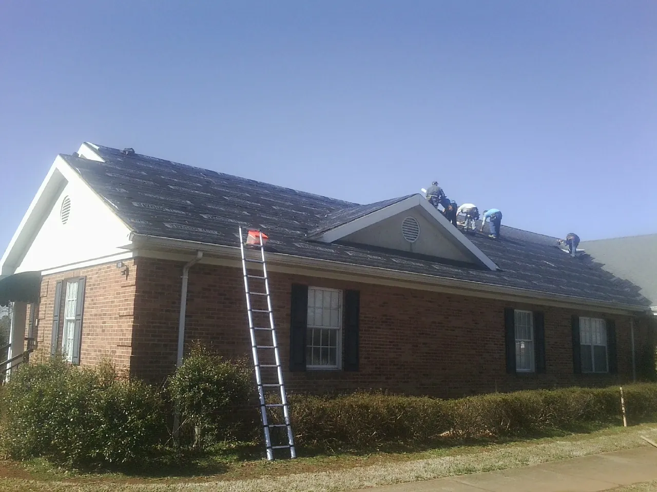 Crew tearing shingles off a brick ranch roof with a ladder set — Metal Roof Repair work by our crew in Alfred