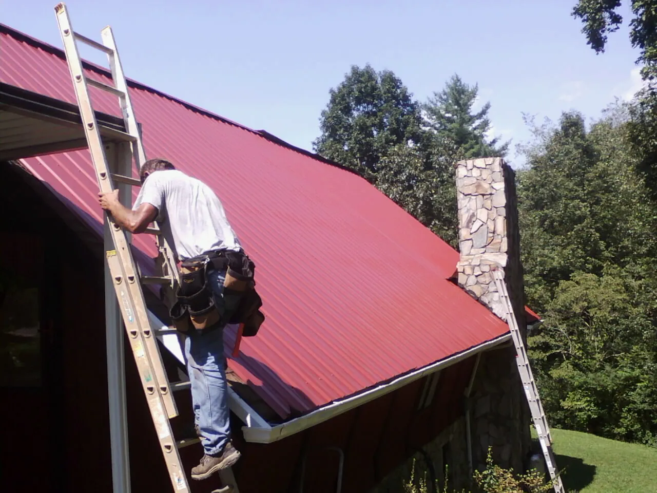 Roofer on a ladder beside a red metal roof and stone chimney — Soffit & Fascia Repair by Roofing Brothers in Alfred