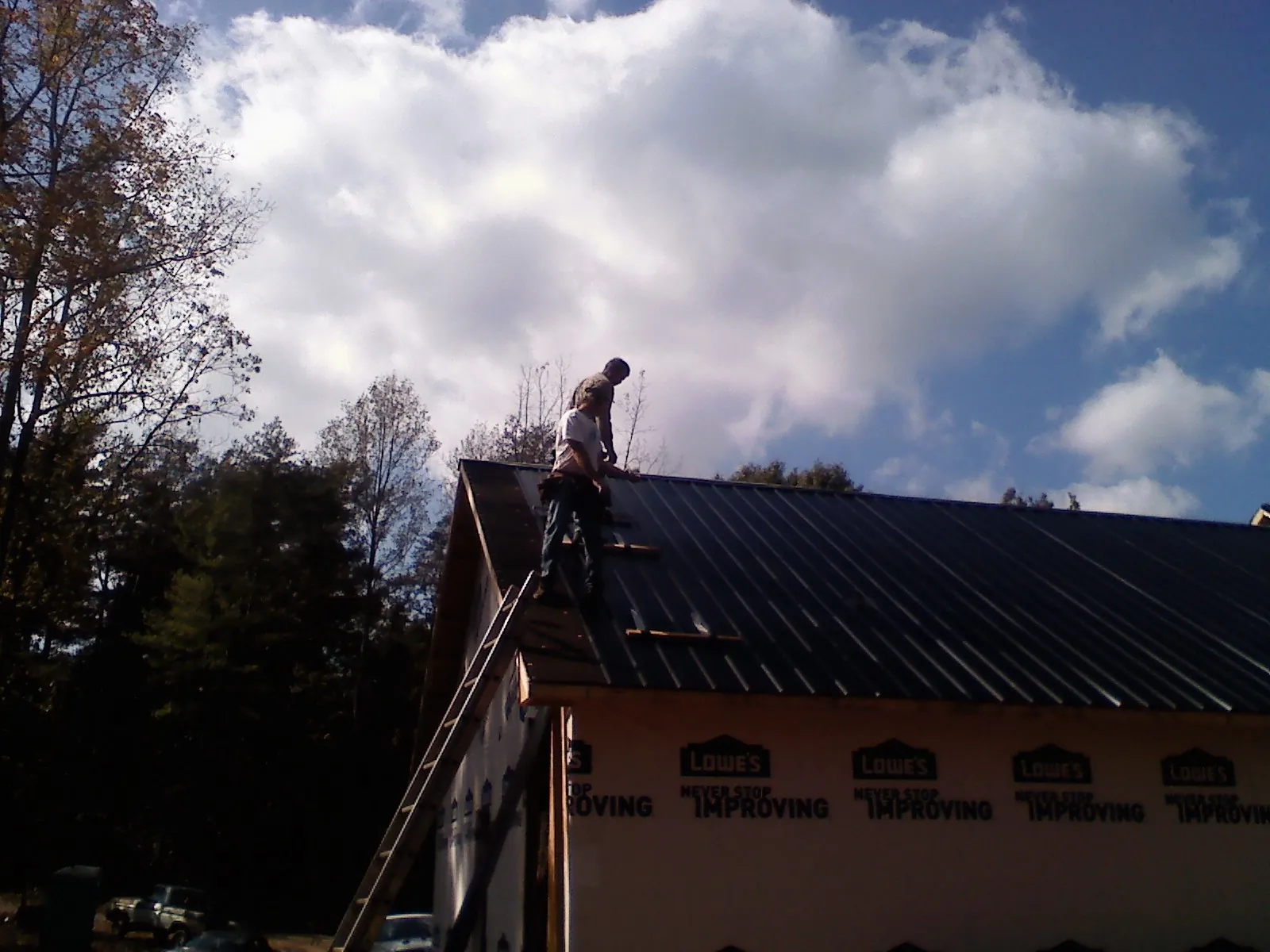 Roofer fastening dark metal panels high against a cloudy sky, from a Roof Inspection job in Alfred